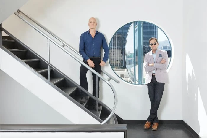 Two men stand on a modern staircase with a circular window overlooking a cityscape. One man wears sunglasses and a blazer.