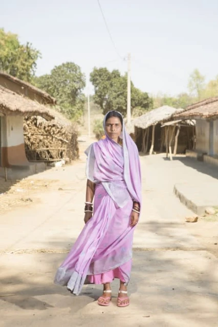 Full shot of adult female wearing a lavender sari with white headscarf standing in rural street with houses and trees in background.