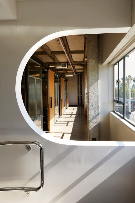 Interior view through an architectural opening reveals a corridor bathed in natural light, with a glimpse of exterior greenery.