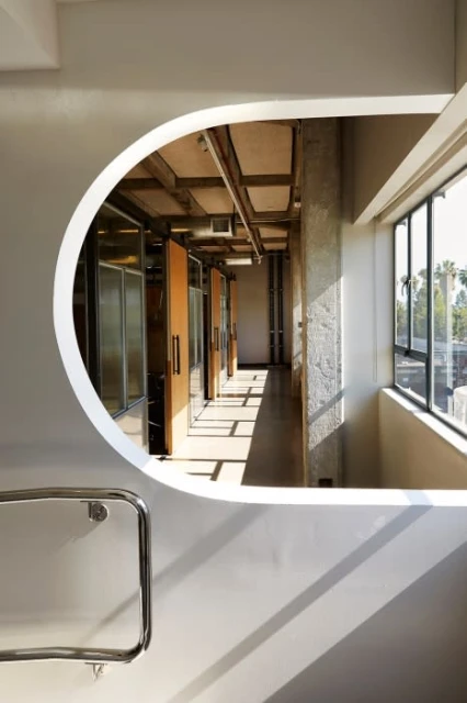 Interior view through an architectural opening reveals a corridor bathed in natural light, with a glimpse of exterior greenery.