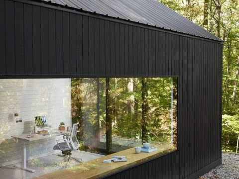 Exterior view of a modern black house with large window showing an office interior with a desk, chair, and laptop in a woodland setting.