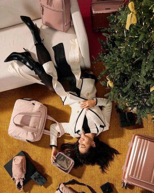 Woman lying on floor surrounded by luggage, Christmas tree, and gifts. Wearing a cream jacket and black boots, she is smiling.