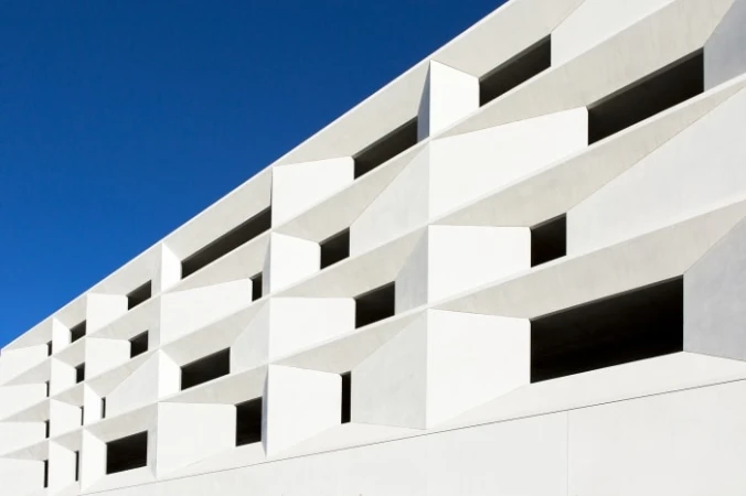 Exterior close-up of a modern, geometric white building with sharp lines, angular design, and repeating window patterns under a vibrant blue sky.