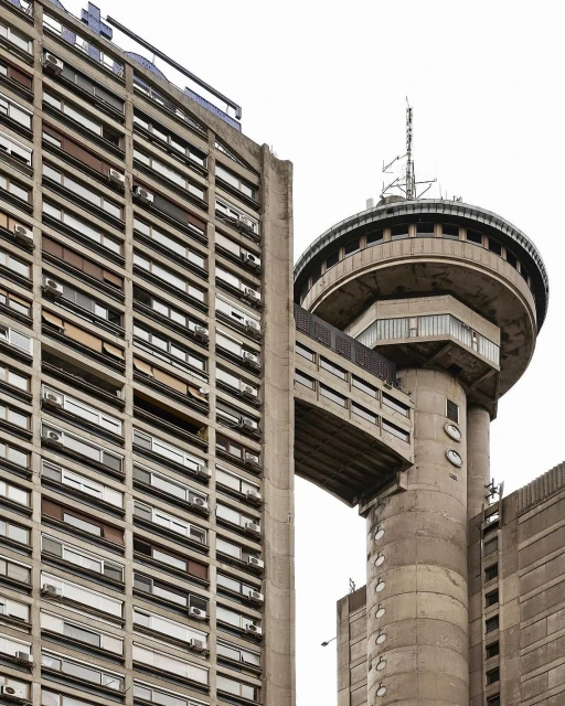 Concrete buildings and a tower are seen against a bright sky. The buildings are modern in style, with repeated window patterns.