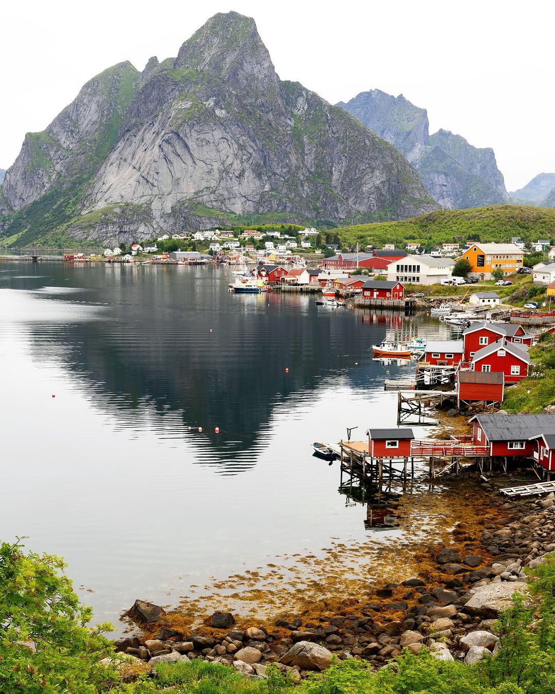 Picturesque coastal village in Norway featuring red houses, boats in water, and towering mountains reflecting on the calm water surface.