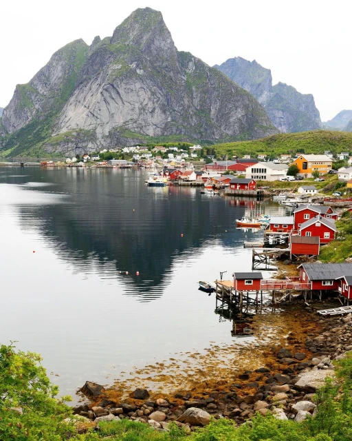 Picturesque coastal village in Norway featuring red houses, boats in water, and towering mountains reflecting on the calm water surface.