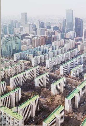 High-angle view of a densely packed urban area featuring rows of mid-rise apartment buildings and skyscrapers in the background, under a hazy sky.