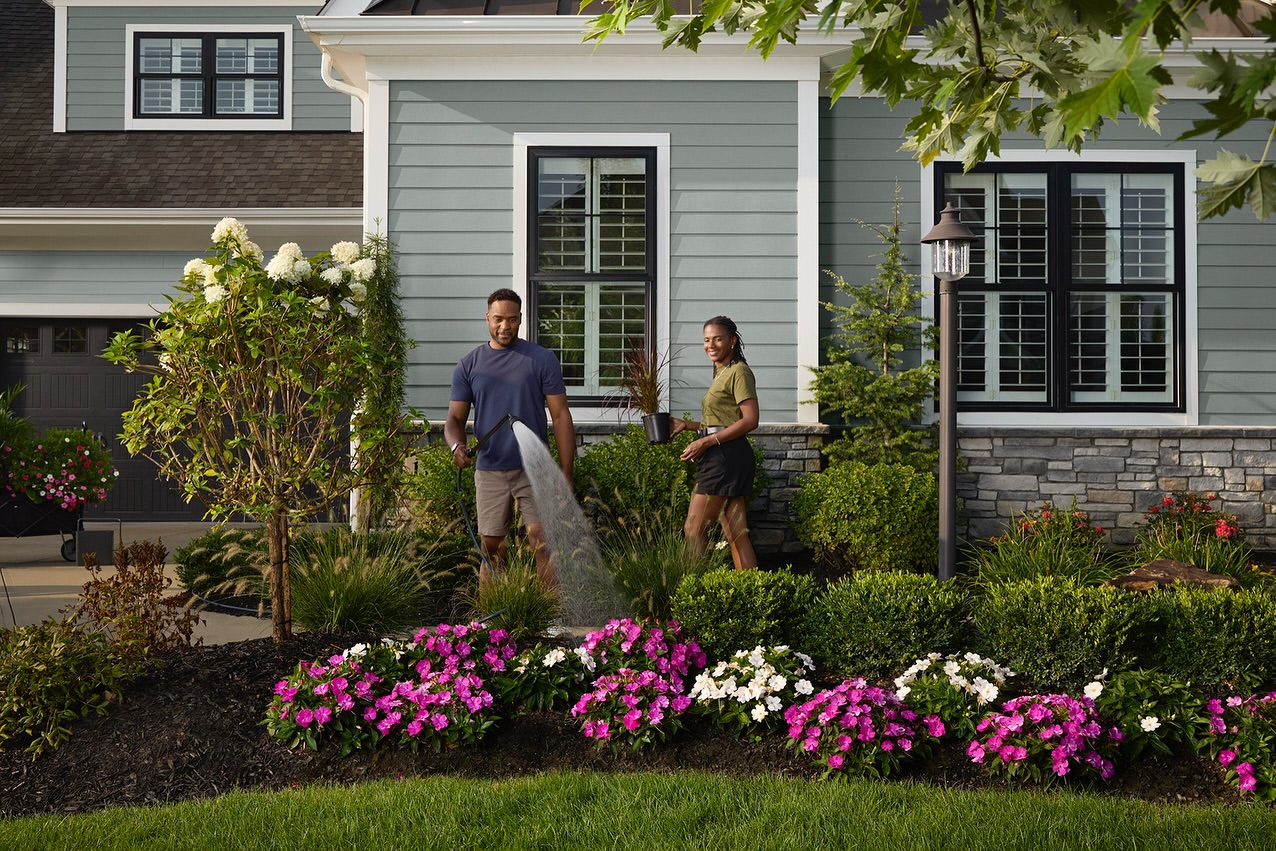 A couple waters flowers in their front yard garden. The man uses a hose while the woman holds a pot with a plant, in daylight.