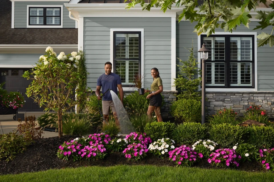 A couple waters flowers in their front yard garden. The man uses a hose while the woman holds a pot with a plant, in daylight.