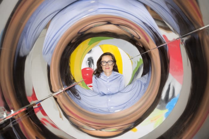 An adult woman with glasses wearing a blue button-down shirt is centered in a distorted mirror tunnel, creating a surreal, abstract effect.