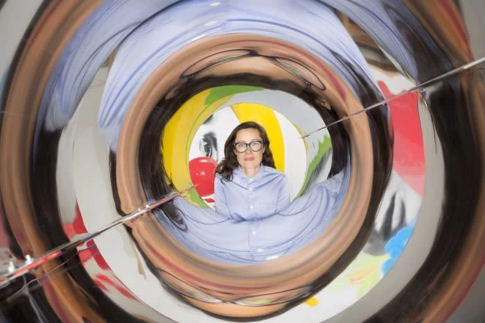 An adult woman with glasses wearing a blue button-down shirt is centered in a distorted mirror tunnel, creating a surreal, abstract effect.