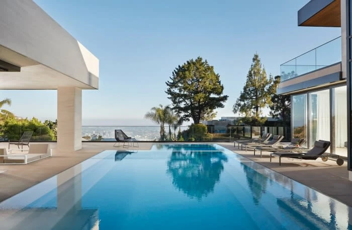 Wide shot of a modern home's outdoor area featuring a large swimming pool reflecting the clear sky and surrounding trees, with lounge chairs.
