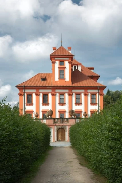 Eye-level view of a symmetrical, orange and white chateau with a red-tiled roof, framed by greenery under a cloudy sky.