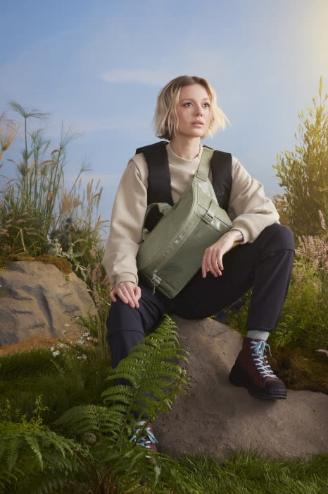 Young woman in nature sitting on rock wearing olive green bag, sweater, and hiking boots against grassy foliage and a blue sky.