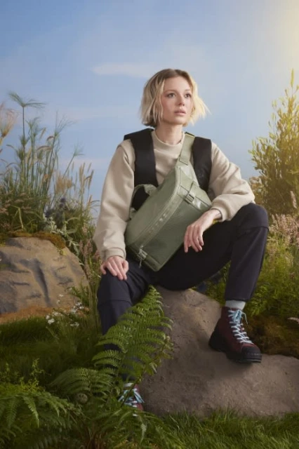 Young woman in nature sitting on rock wearing olive green bag, sweater, and hiking boots against grassy foliage and a blue sky.