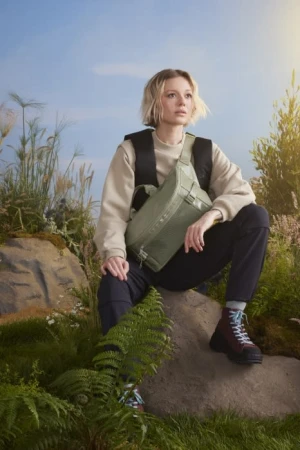 Young woman in nature sitting on rock wearing olive green bag, sweater, and hiking boots against grassy foliage and a blue sky.