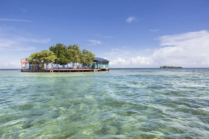 Island getaway featuring a small wooden house surrounded by trees, sitting on clear, turquoise water under a bright blue sky.