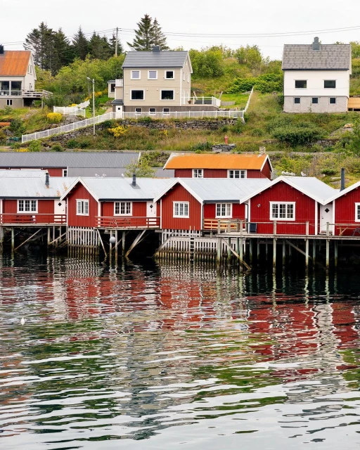 Scenic view of waterfront red wooden houses in Norway with reflections on the water and lush green hillsides under a cloudy sky.