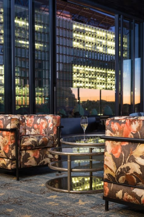 Cozy seating area with floral armchairs, a round glass table, and a view of a lit urban building seen through a large window.