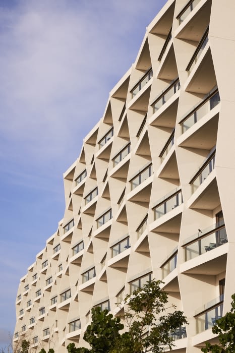 Exterior view of modern architecture with geometric facade, balconies, and angular design against a partly cloudy blue sky and green trees.