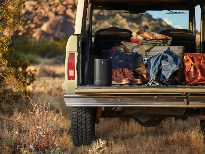 Portable speaker and outdoor gear, including a cooler, blanket, bags, and boots, are in the back of a SUV amid a natural desert setting.