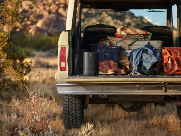 Portable speaker and outdoor gear, including a cooler, blanket, bags, and boots, are in the back of a SUV amid a natural desert setting.