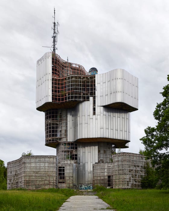 Futuristic brutalist monument with antenna, metallic cladding, open grid areas, overgrown landscape in overcast, daytime. Architecture.