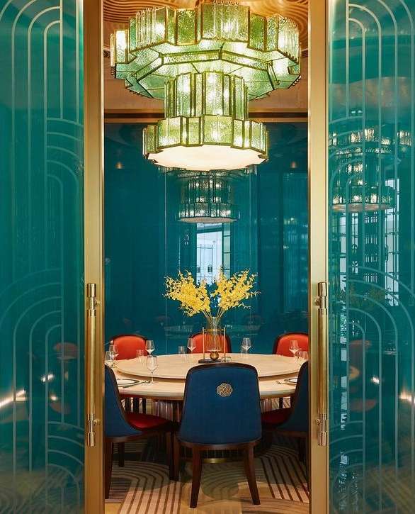 Ornate light fixture illuminates a dining table set with clear glassware and surrounded by colorful chairs in a restaurant setting.
