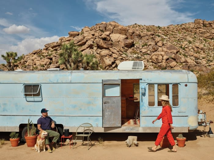 A light blue vintage travel trailer in a desert setting with a man, a woman in red, and a dog. Rocky hills and blue skies.