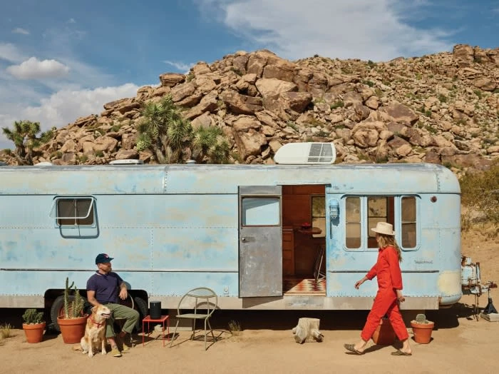 A light blue vintage travel trailer in a desert setting with a man, a woman in red, and a dog. Rocky hills and blue skies.