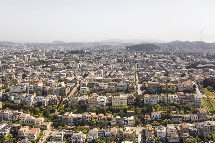 Aerial view of San Francisco cityscape featuring rows of houses, skyscrapers, roads, and green parks under a clear sky.