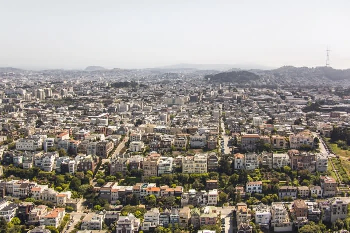 Aerial view of San Francisco cityscape featuring rows of houses, skyscrapers, roads, and green parks under a clear sky.