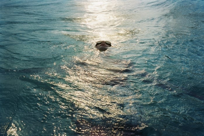 Person with swimming goggles floating in a sun-drenched body of turquoise water, rippling across the frame from above eye-level.