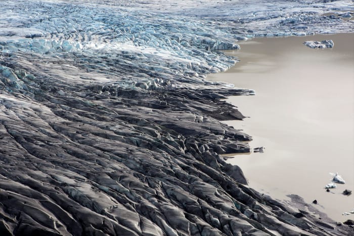 Aerial view of a rugged glacier landscape meeting the edge of a still, light brown glacial lake in a remote and icy environment.