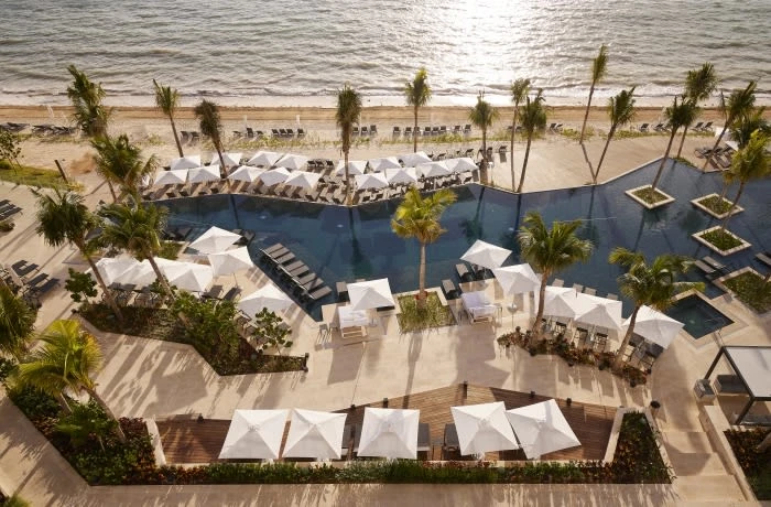 High-angle view of a luxury resort with palm trees, a pool with square islands, and white umbrellas along a sandy beach by the ocean.