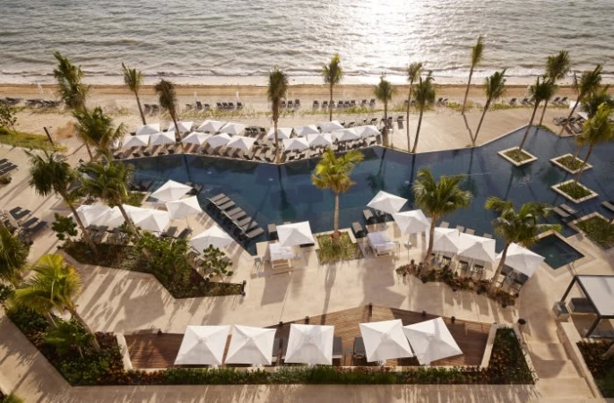 High-angle view of a luxury resort with palm trees, a pool with square islands, and white umbrellas along a sandy beach by the ocean.