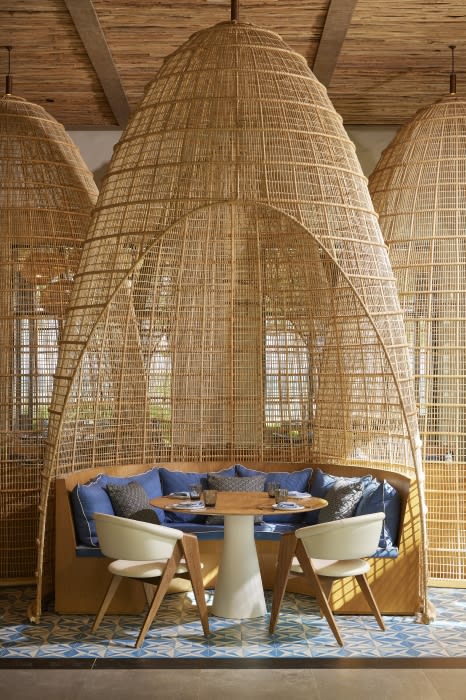 Interior of a restaurant with a round table, white chairs, blue pillows, and natural woven booths under a textured ceiling.