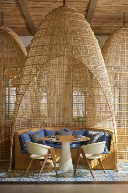 Interior of a restaurant with a round table, white chairs, blue pillows, and natural woven booths under a textured ceiling.