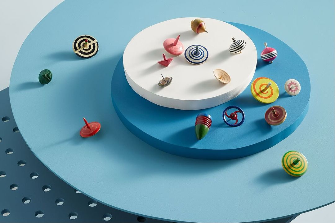 Overhead shot displays a variety of colorful wooden spinning tops arranged on white and blue circular platforms against a blue background.
