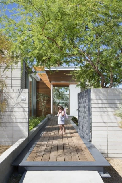 Young girl in white dress stands on wooden walkway near modern home exterior with green tree, concrete walls and metal gate.