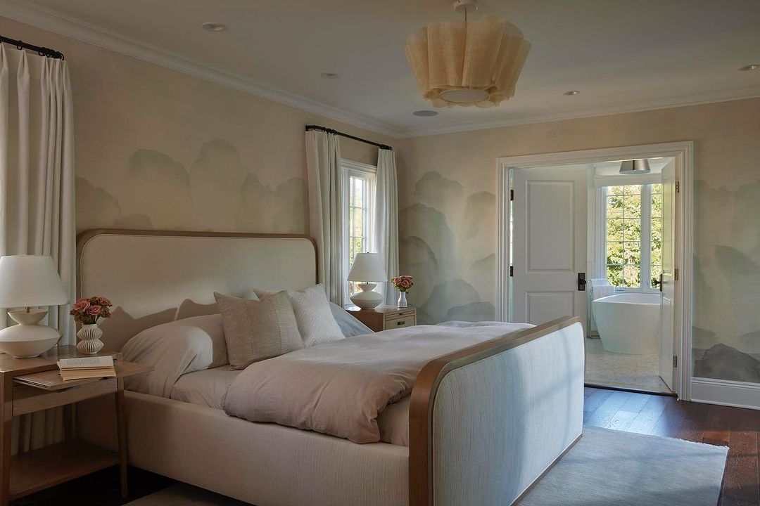Bedroom interior featuring a bed with neutral linens, bedside tables with lamps, and light-colored walls with muted landscape design.
