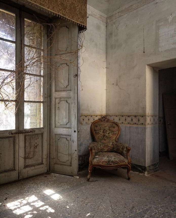 Old floral armchair positioned inside an abandoned room with aged walls and a large window covered in vines and bright sunlight.