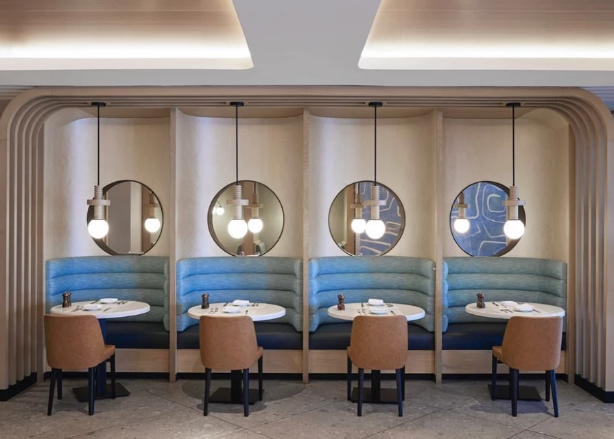 Restaurant dining booths with upholstered seating, round tables, pendant lights, and mirrored wall details in a modern interior space.