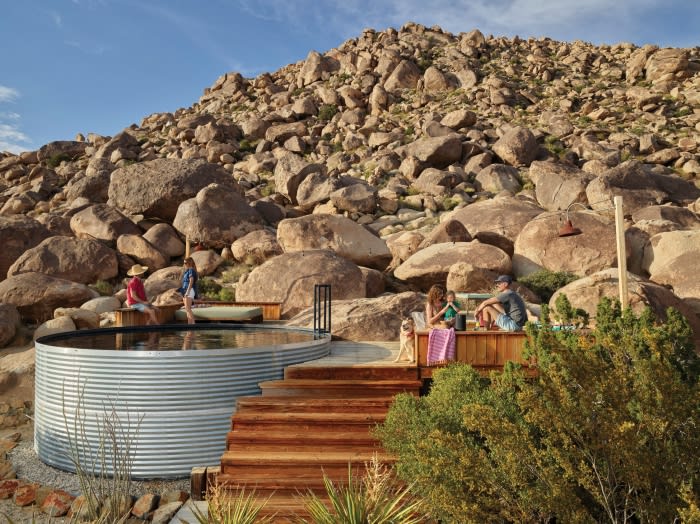 Outdoor gathering with people relaxing on a wooden deck and benches next to a metal-sided stock tank pool, surrounded by rocky hills.