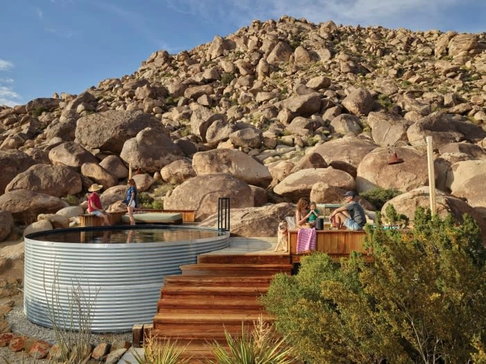 Outdoor gathering with people relaxing on a wooden deck and benches next to a metal-sided stock tank pool, surrounded by rocky hills.