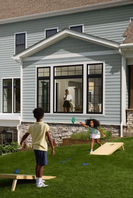 Two children play cornhole on a green lawn in front of a light blue house with black-framed windows, while an adult stands inside.