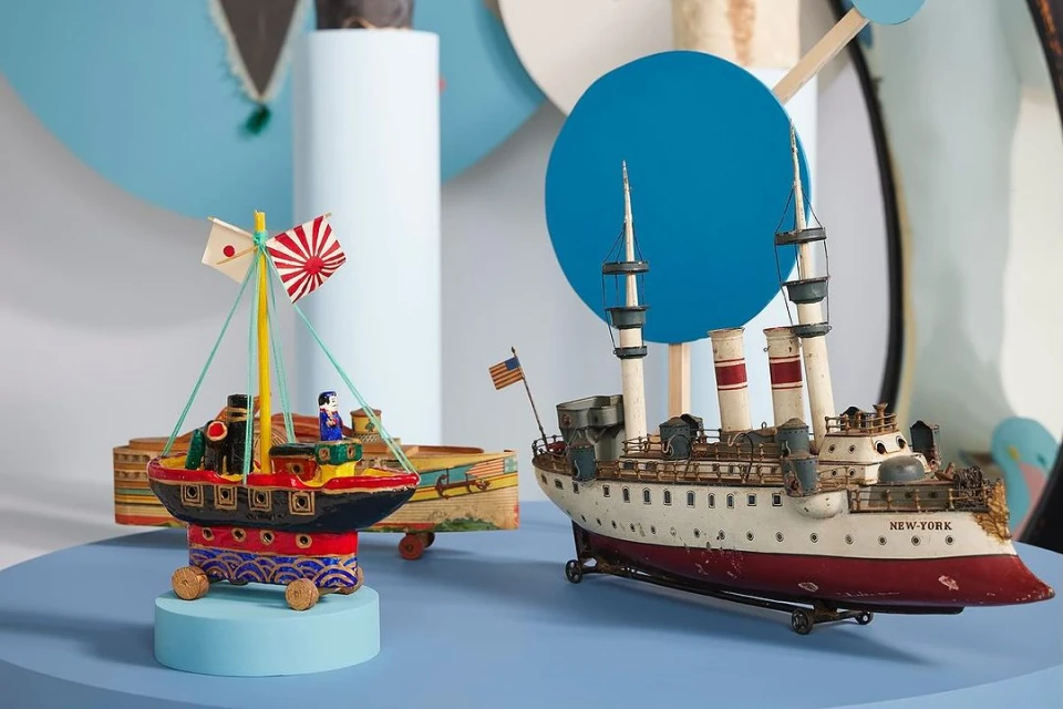 Vintage toy boats, one with Japanese flag and one labeled New York, displayed on blue surface with geometric white and blue background.