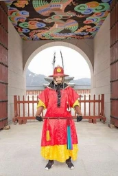 A guard in traditional red and yellow military robes and helmet stands centered under an ornate gateway arch.
