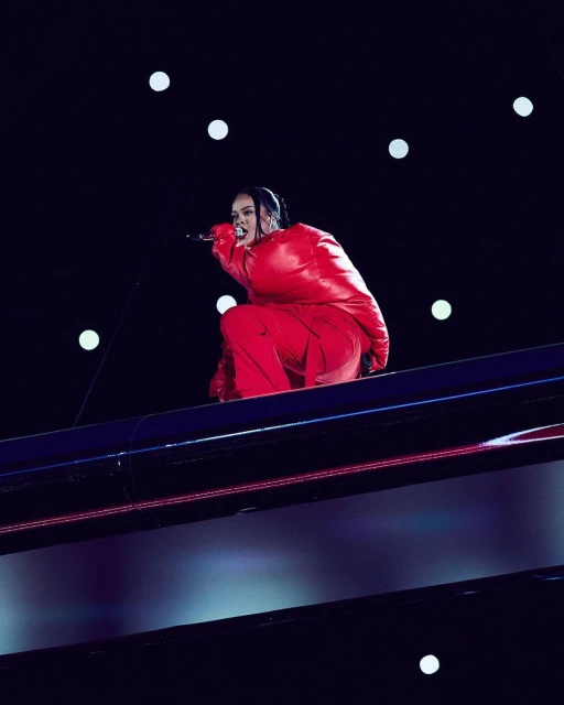 Singer Rihanna performs in a red outfit on a high platform under bright lights during a performance at night.