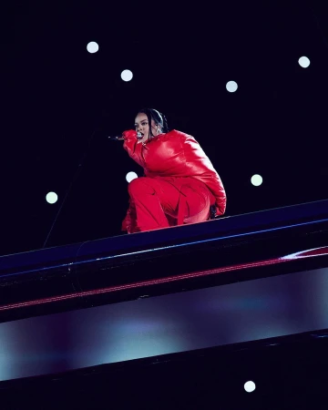 Singer Rihanna performs in a red outfit on a high platform under bright lights during a performance at night.